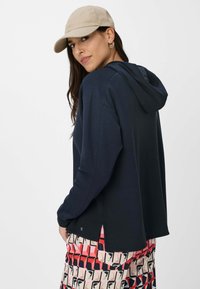 Felpa con cappuccio blu navy dal texture morbido; presenta fessure laterali e un logo. Abbinata a pantaloni corti a fantasia nei toni del rosso, rosa e crema.