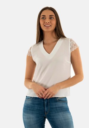 Vit blus med korta ärmar som har spetsdetaljer, V-ringning och en slät textur; kombinerad med blå denimjeans.