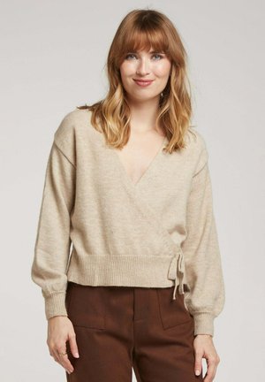 Pull en beige à enrouler avec un col en V, ourlet côtelé et manches longues. Texture douce ; présente un lien à la taille. Associé à un pantalon marron.