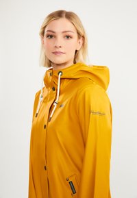 Chaqueta de lluvia impermeable amarilla con capucha, que cuenta con cordones blancos, botones a presión y una marca en la manga. Textura suave y brillante.