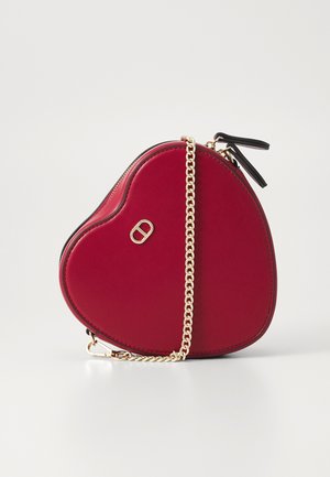 Sac à main rouge en forme de cœur avec bandoulière chaîne dorée et petit logo doré à l'avant, sur fond blanc uni.