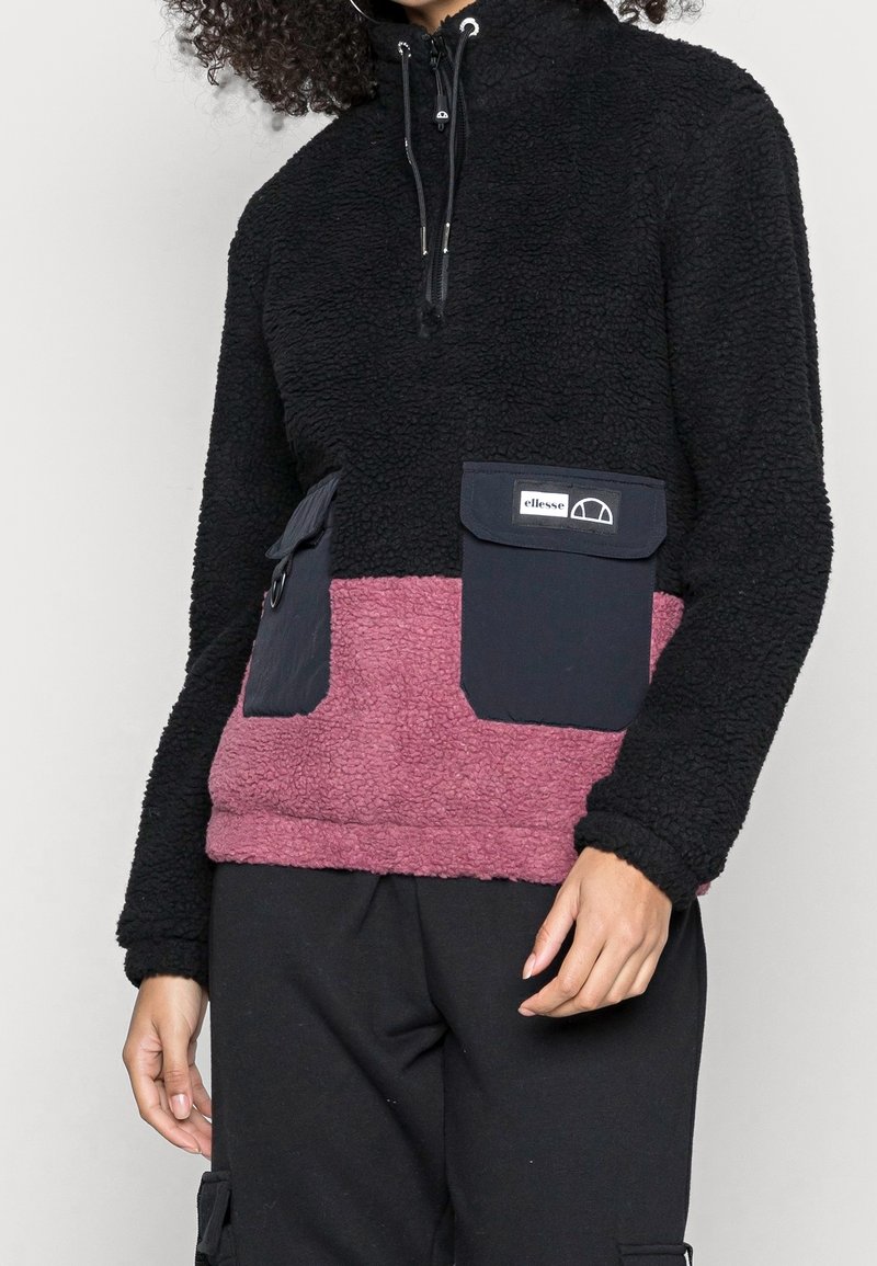 Personne portant un pull polaire noir et mauve avec de grandes poches avant et un col à zip quart, associé à un pantalon noir.