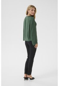 Haut vert à manches longues avec une coupe décontractée, fabriqué en tissu léger. Associé à un jean skinny noir et des mules noires à talons.