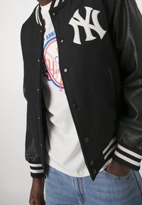 Person som bär en varsityjacka med svarta läderslag och vit NY-logga samt en vit t-shirt med grafiskt tryck i blått och rött, ihop med blå jeans.