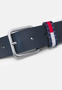 Cinturón de cuero azul marino con una hebilla rectangular de plata. Presenta una banda trenzada con rayas rojas, blancas y azules y el logo "TOMMY JEANS".
