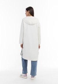 Lange witte regenjas met een capuchon, met een gladde, glanzende textuur. In combinatie met blauwe spijkerbroeken en meerkleurige sneakers met een dikke zool.