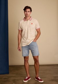 Crème polo shirt met roodgestreepte kraag en mouwen, versierd met een subtiele bladvorm. Gecombineerd met lichte denimshorts en bordeaux sneakers.