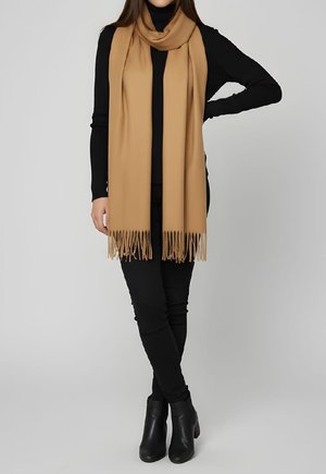 Mujer con cuello de tortuga negro, pantalones negros, botines negros y una bufanda larga de color camel con flecos, de pie frente a un fondo liso.