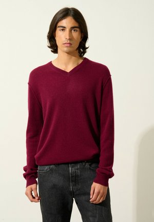 Pull en V bordeaux en tissu doux, présentant des manches longues avec des poignets côtelés, associé à un jean noir texturé.