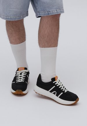 Czarne sportowe sneakersy z białymi akcentami, górna część z materiału, gumowa podeszwa, pomarańczowa metka marki; noszone z białymi, prążkowanymi skarpetami i dżinsowymi szortami.