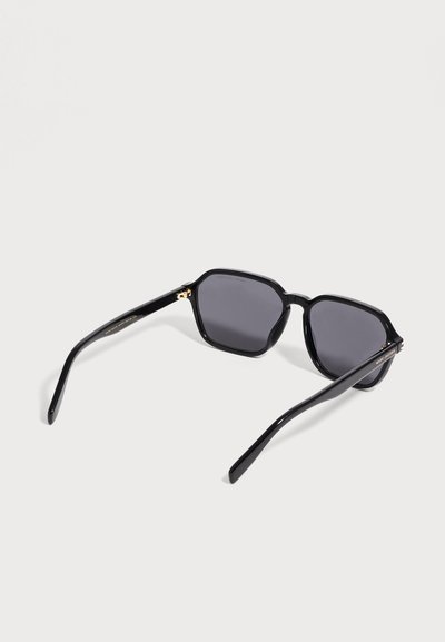 Lunettes de soleil noires à monture carrée avec verres foncés et branches fines, exposées sur une surface gris clair.
