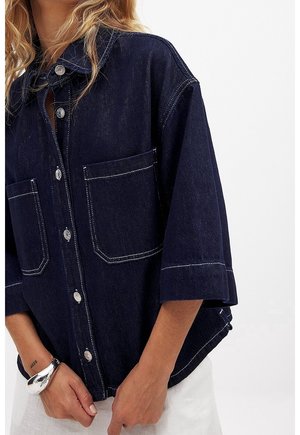 Vrouw die een donker denim blouse met grote voorzakken en witte stiksels draagt, gecombineerd met een witte broek en een zilveren armband.