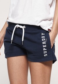 Frau trägt marineblaue Superdry-Shorts mit weißer Kordel und weißem T-Shirt, Hände in den Taschen.