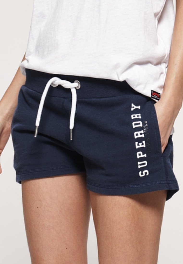 Frau trägt marineblaue Superdry-Shorts mit weißer Kordel und weißem T-Shirt, Hände in den Taschen.