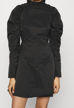 Kobieta ubrana w czarną sukienkę z marszczonymi, bufkiowymi długimi rękawami i wysokim dekoltem, stojąca na tle jednolitego, jasnego tła.