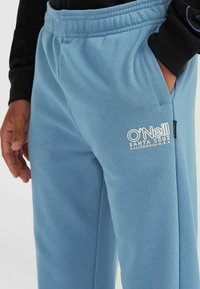 Helle blaue Jogginghose aus weichem Material, mit einem elastischen Bund, Seitentaschen und einem weißen gestickten Logo am Oberschenkel.