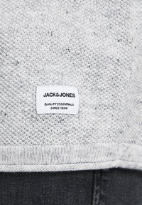 Pull en maille grise avec une étiquette rectangulaire blanche indiquant "JACK & JONES QUALITY ESSENTIALS SINCE 1990" au-dessus d'un pantalon noir.