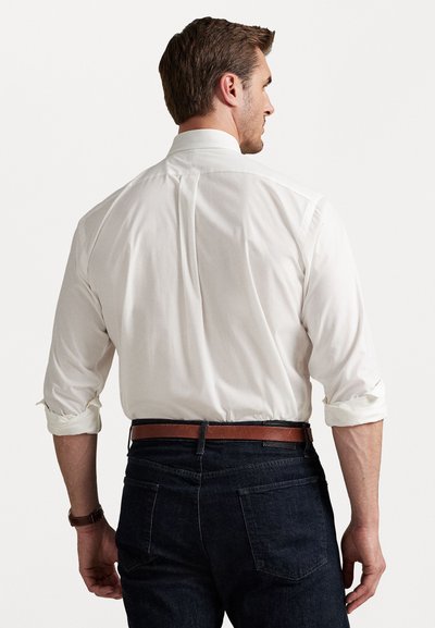 Chemise blanche à manches retroussées, assortie à un jean en denim foncé et une ceinture en cuir marron. La vue arrière met en valeur l'ajustement et la texture de la chemise.