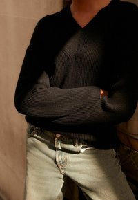 Pull noir en maille côtelée avec col en V, associé à un jean en denim clair. Le pull présente une coupe décontractée et une surface texturée.
