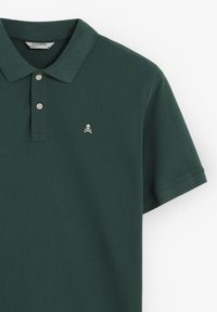 Polo en color verde oscuro hecho de algodón, con una abertura de tres botones, mangas cortas y un logo de calavera y tibias cruzadas en blanco en el pecho.