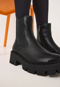 Bottes de cheville en cuir noir avec des panneaux latéraux en élastique texturé et des semelles épaisses et à rainures. Fini lisse et éclat subtil. Détail de fermeture éclair sur un côté.