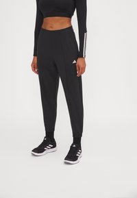 Zwarte sportieve joggers met een tapered fit, met het witte Adidas-logo en drie strepen langs de zijkanten. Gepaard met zwarte sportschoenen.