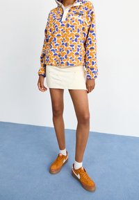 Fleece pullover dengan pola abstrak oranye, ungu, dan krim; kerah setengah resleting; dipadukan dengan rok mini putih dan sepatu sneakers oranye.