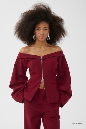 Γυναίκα με πυκνά, σγουρά μαλλιά, που φοράει μπορντό μπλούζα με ανοιχτούς ώμους και ασημένιο φερμουάρ, καθώς και παντελόνι που ταιριάζει, μπροστά σε απλό φόντο.