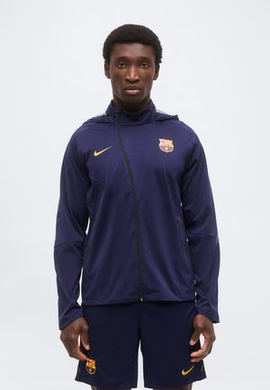 Homme portant une veste et un short bleu marine du FC Barcelone avec des logos Nike jaunes, debout devant un fond clair uni.