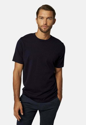 Uomo con capelli castano chiaro e barba, che indossa una maglietta nera a maniche corte semplice e pantaloni scuri su sfondo bianco.