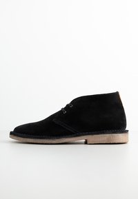Stivaletti alla caviglia in suede nero con punta rotonda, cuciture a contrasto e un accento in pelle color cuoio sul tallone. Presentano una suola piatta in gomma.