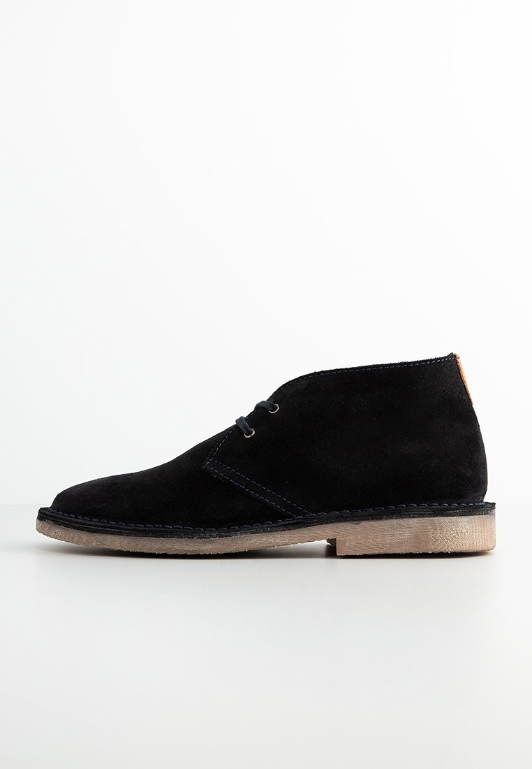 Stivaletti alla caviglia in suede nero con punta rotonda, cuciture a contrasto e un accento in pelle color cuoio sul tallone. Presentano una suola piatta in gomma.
