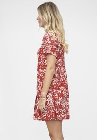 Punainen kukkakuosinen mekko, jossa on lyhyet hihat, kokoontaitettu vyötärö ja valkoiset kukkakuviot. Sileä kangas ja liehuva helmareunus.