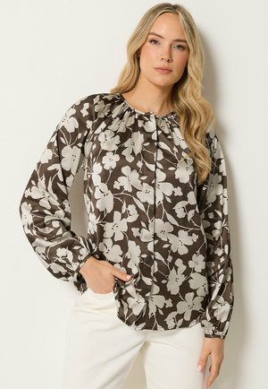 Vrouw met lang blond haar die een zwart-wit bloemenprint blouse met lange mouwen en een witte broek draagt, staand met één hand in de zak.