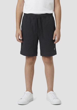 Kind draagt gestructureerde zwarte short met trekkoord, witte sneakers en een wit T-shirt, staand tegen een effen grijze achtergrond.