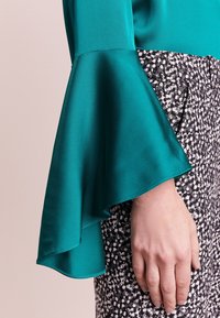 Teal satynowa bluzka z rozpierzonymi rękawami połączona z czarno-białą spódnicą w wzory. Widoczne gładka tekstura i płynne detale projektu.