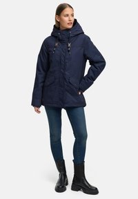 Mörkblå vadderad jacka med huva, dragkedja framtill, två stora fickor och justerbara dragskor. Bärs med blå jeans och svarta kängor.