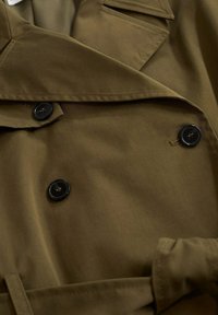 Oliven grøn trenchcoat detalje med sorte knapper, tekstureret stof, dobbeltbrystet design og bælteaccent.