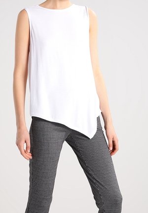 Ärmelloses asymmetrisches weißes Top, kombiniert mit schwarz-weiß karierten Slim-Fit-Hosen an einer stehenden Person.