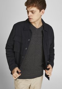 Mörkblå knappad jacka med två bröstfickor över en mörkgrå V-halsad tröja; modellen bär ljusbeiga byxor och justerar jackan med händerna.