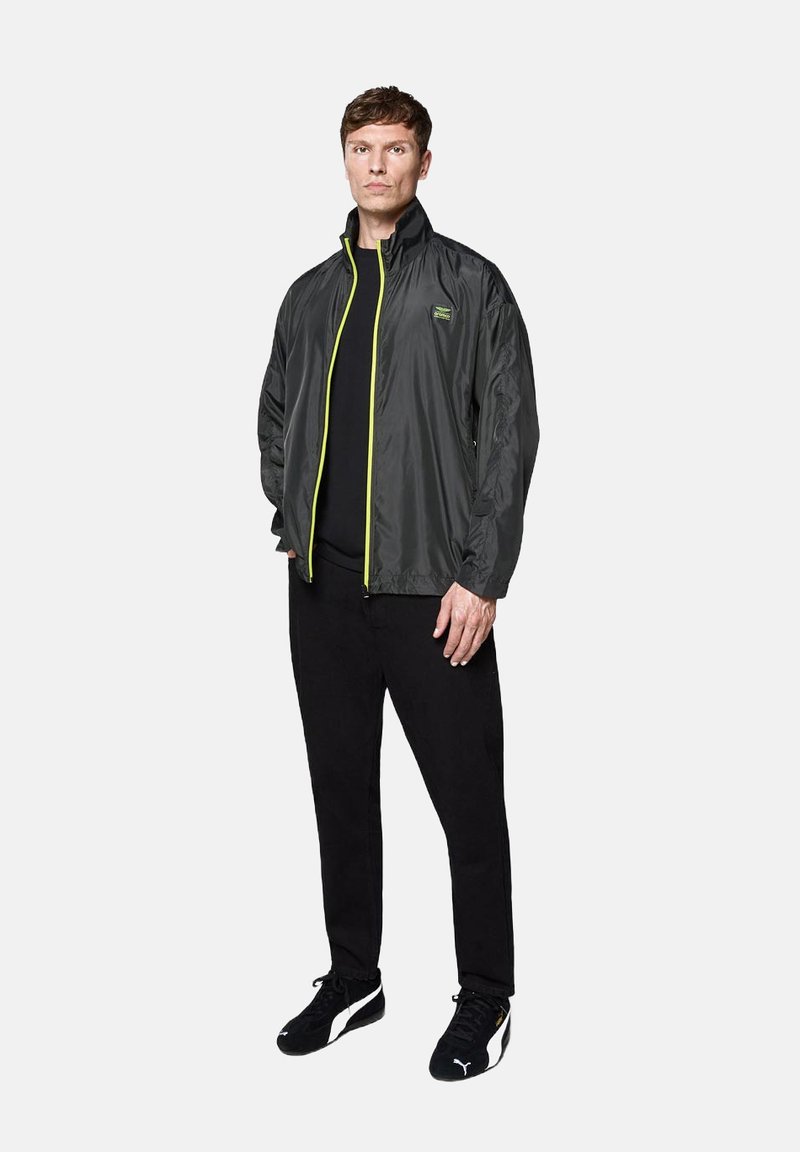 Lekka, szara kurtka z wysokim kołnierzem i neonowo-żółtym zamkiem. Noszona na czarną koszulę, w zestawieniu z czarnymi spodniami i czarnymi sneakersami.