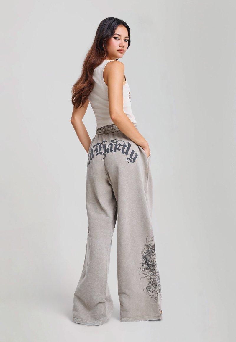 Woman with long dark hair wears white sleeveless top and gray wide-leg pants with black text and dragon design, standing with hands in pockets.