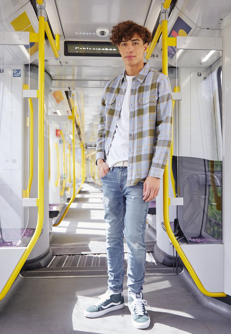 Camisa de cuadros en azul claro y verde, camiseta blanca, jeans rasgados azul claro y zapatillas verdes, de pie en un pasillo de tren.