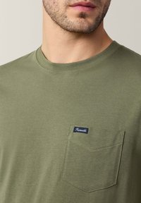 Koszulka z bawełny w oliwkowym kolorze z okrągłym dekoltem, krótkimi rękawami i kieszonką na piersi z niebieską metką. Gładka tekstura, casualowy design.