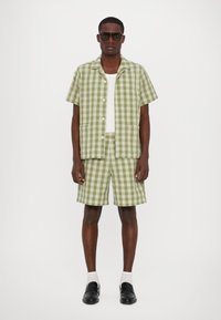 Πράσινο και λευκό καρώ κοντομάνικο πουκάμισο με αντίστοιχες σορτς, φτιαγμένο από ελαφρύ ύφασμα. Συνδυασμένο με μαύρα παπούτσια και μεγάλα γυαλιά ηλίου.