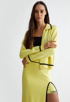 Cardigan giallo a maglia con dettagli neri e zip, abbinato a una gonna aderente coordinata con spacco laterale e dettagli con logo.