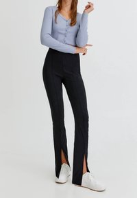 Cardigan largo de manga larga en azul claro y pantalones ajustados negros con aberturas laterales, combinados con zapatillas blancas. Textura suave y silueta ajustada.