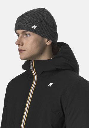 Berretto grigio a coste indossato da una persona, abbinato a una giacca nera con cerniera multicolore e un logo sul petto. Primo piano del profilo laterale.