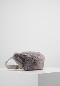 Kulatá šedá taška z umělé kožešiny s jemnou strukturou. Obsahuje stříbrný zip a béžový nastavovací popruh pro bezpečné nošení.
