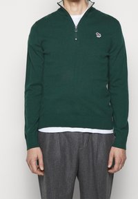 Maglione zip-up verde scuro con maniche lunghe, texture a maglia e piccolo logo ricamato sul petto, indossato sopra una camicia bianca.